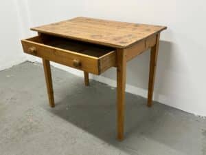 Wooden table with a single open drawer, set against a plain wall on a concrete floor.