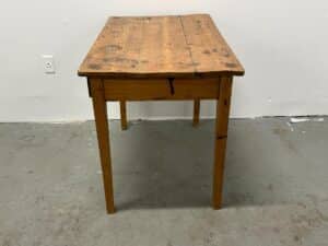 A rustic, vintage wooden table with a weathered surface and four legs, placed against a plain white wall.