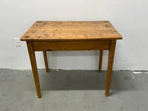 A rustic wooden table with visible wear, positioned against a plain white wall on a cement floor.
