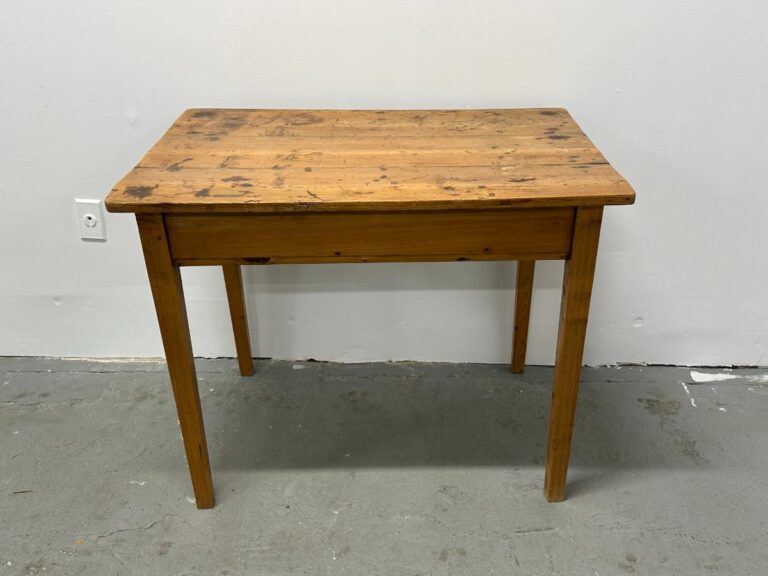 A rustic wooden table with visible wear, positioned against a plain white wall on a cement floor.
