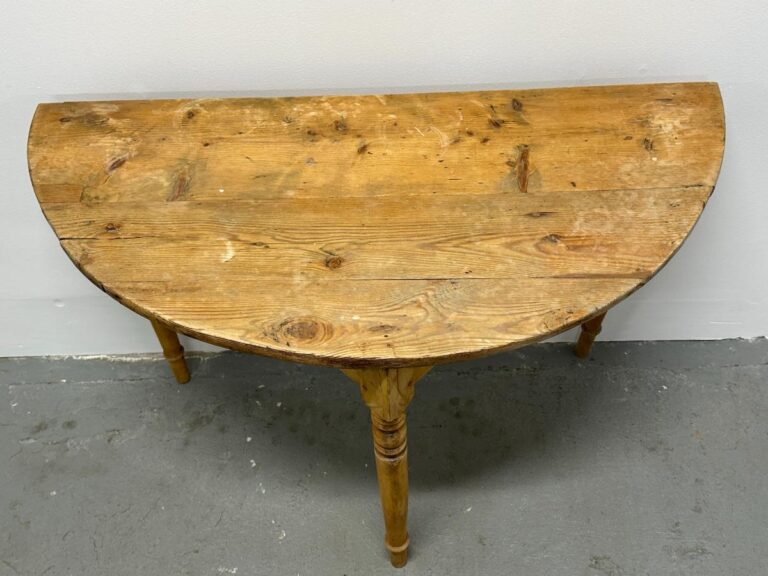 Half-moon shaped wooden table with scuffed surface and three turned legs against a plain wall.