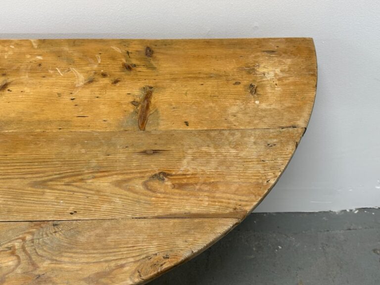 Close-up of a round wooden table with visible grain and knots, against a light gray wall and floor.