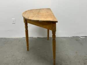A half-circle wooden table with four turned legs against a white wall on a concrete floor.