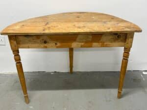 Wooden half-moon table with two turned legs against a white wall on a gray floor.