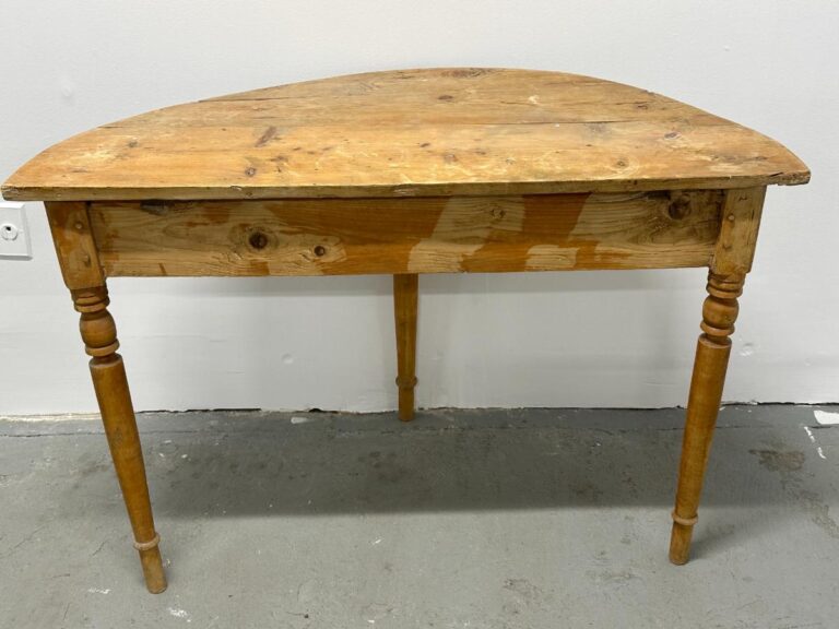 Wooden half-moon table with two turned legs against a white wall on a gray floor.