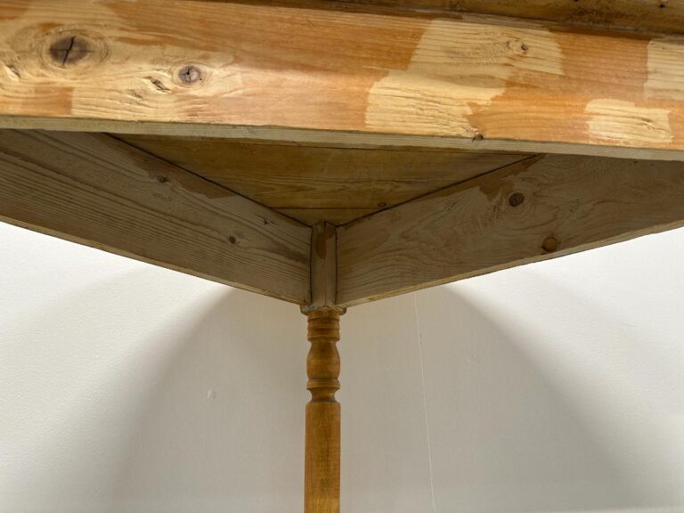 Underneath view of a wooden table corner with one turned leg and visible wood grain patterns.