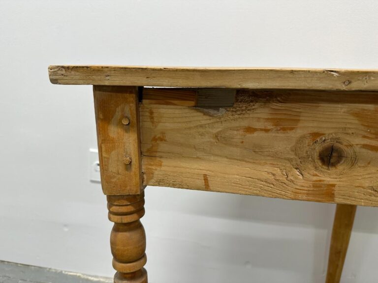 A wooden table with a rough, rustic design and a visible knot hole in the leg against a plain wall.