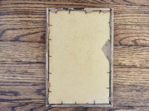 The worn back of an antique framed VC Andrews "Nobody Loves Me" print, copyright 1906 by Charles Scribner & Sons, shows peeling brown paper as it lays on a wooden floor.