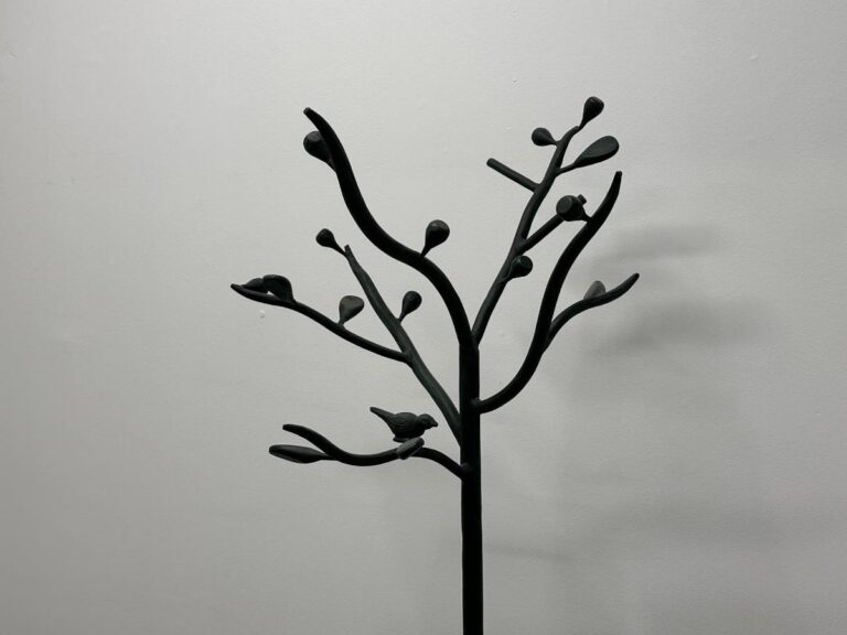Sculptural coat rack resembling a tree with branches and a bird, set against a plain, light-colored wall.