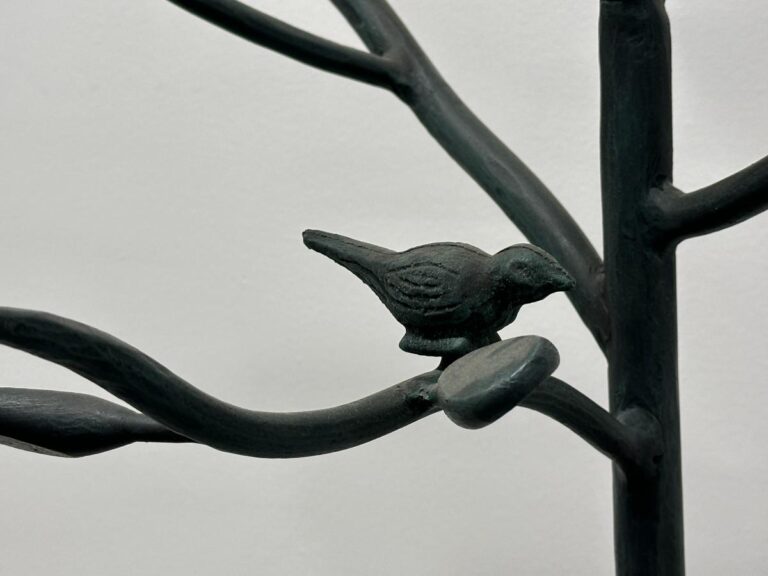 Metal bird sculpture perched on a branch, with a white background.