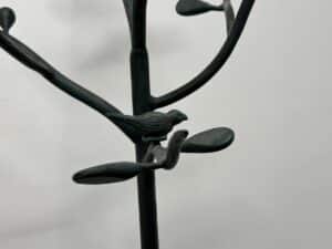 Metal sculpture of a bird perched on a stylized branch with leaves against a plain background.