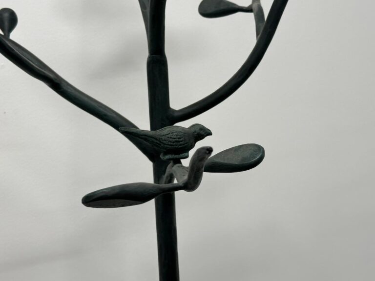 Metal sculpture of a bird perched on a stylized branch with leaves against a plain background.