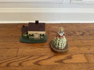 A cast iron door stop set features a painted Southern lady in a dress and a Cape Cod cottage on a wooden floor.
