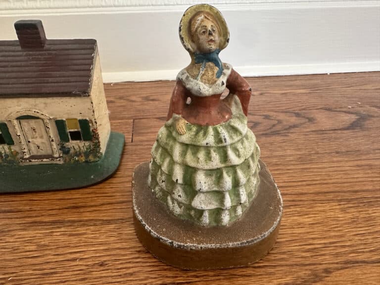 A Cast Iron Painted Southern Lady in a ruffled dress and bonnet stands beside a Cape Cod Cottage Door Stop on a wooden floor.