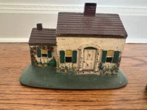 A small, rustic ceramic house with green shutters on a wooden surface; product name: Cast Iron Painted Southern Lady & Cape Cod Cottage Door Stops.