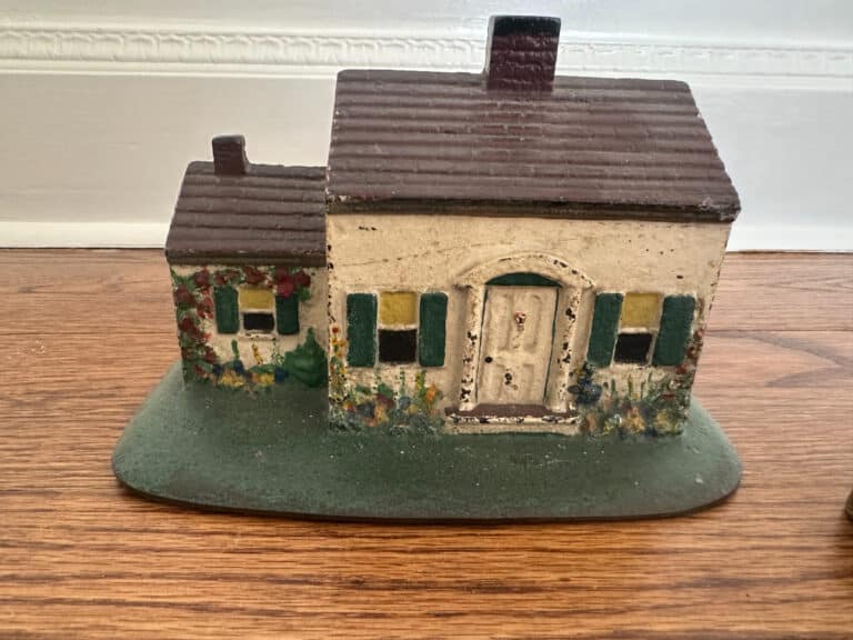 A small, rustic ceramic house with green shutters on a wooden surface; product name: Cast Iron Painted Southern Lady & Cape Cod Cottage Door Stops.