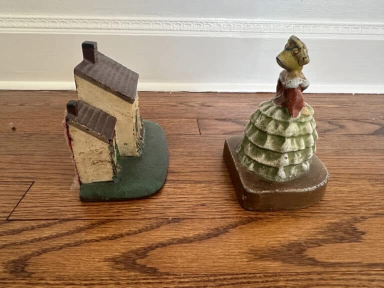 Two cast iron door stops on a wooden floor: one shaped like a Southern lady in a ruffled dress, the other like a Cape Cod cottage.