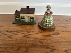 A cast iron painted Southern lady figurine and a Cape Cod cottage door stop on a wooden floor.