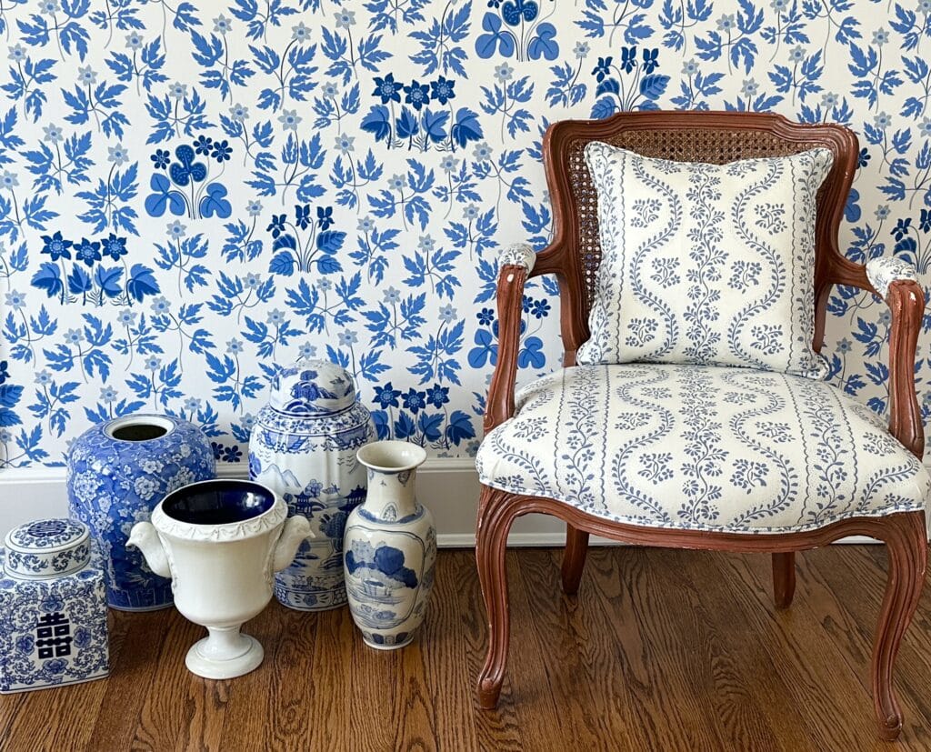 Ornate chair with blue floral cushion beside blue and white pottery against a floral wallpaper background.