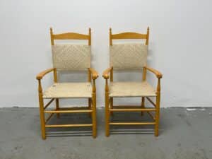 Two wooden chairs with woven seats and backs, positioned side by side against a plain white wall.