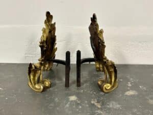 Ornate gold and bronze decorative andirons on a gray surface against a light wall.