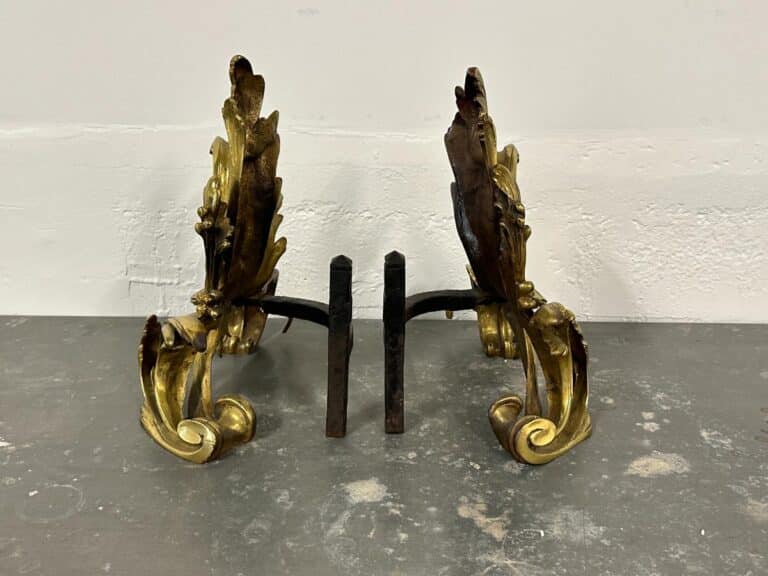 Ornate gold and bronze decorative andirons on a gray surface against a light wall.