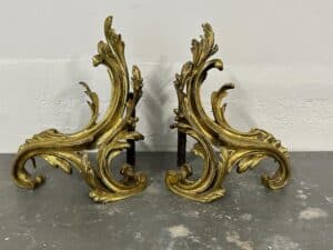 Two ornate, gold-colored fireplace andirons with swirling leaf designs on a gray surface.