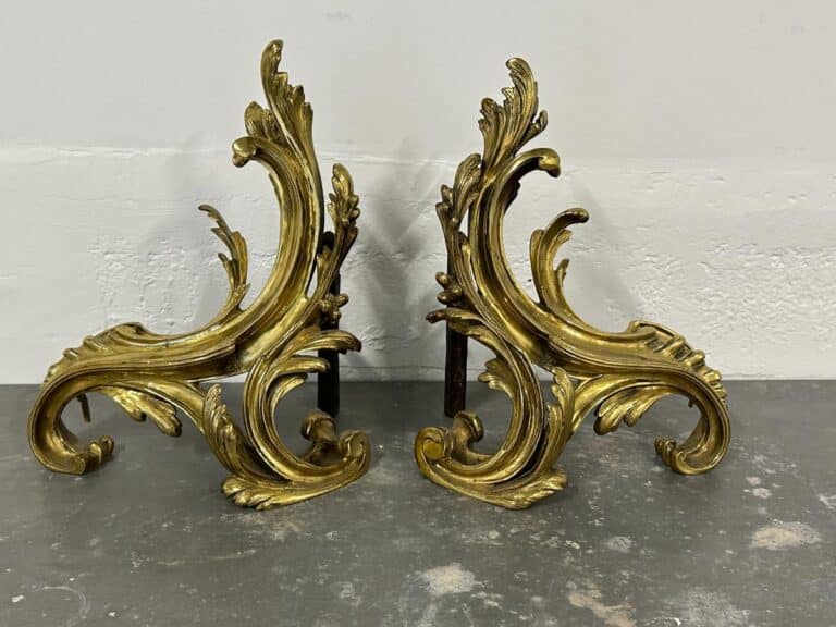 Two ornate, gold-colored fireplace andirons with swirling leaf designs on a gray surface.