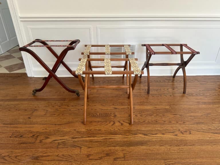 Three luggage racks—Antique Regency, Vintage Scheibe, and Reeded Leg—from the set of four featuring patterned fabric straps on a hardwood floor against a white-paneled wall.