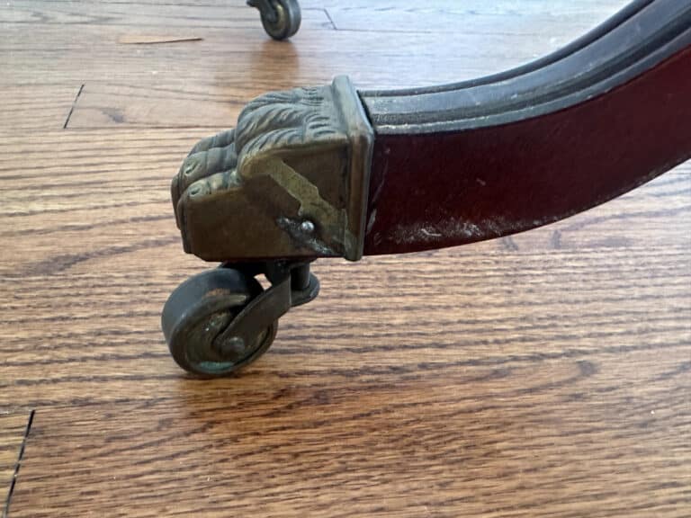 Close-up of a French Carved Mahogany Vanity Ladies Desk leg featuring an ornate metal caster on a hardwood floor.