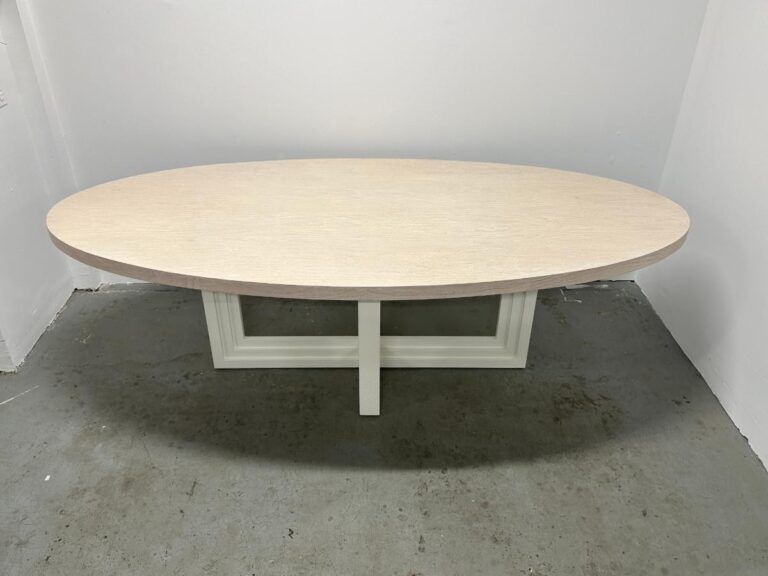 The Mr. Brown London Florence Dining Table, featuring an oval wooden top and modern geometric base, sits elegantly in a plain white-walled room.