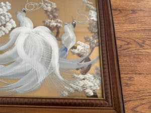 The "Birds of Paradise" painting, featuring two white peacocks with long tails surrounded by trees, is signed lower right and displayed in a wooden frame on a wooden floor.