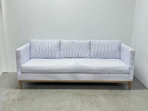 A Serena & Lily Barton Sofa in light blue with stripes and wooden legs sits against a plain white wall.