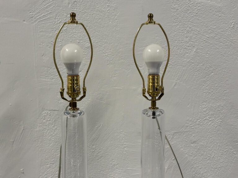 Two Simon Pearce Glass Column Lamps, sold as a pair, featuring clear glass and brass fixtures with unlit white bulbs, sit against a textured wall without shades.