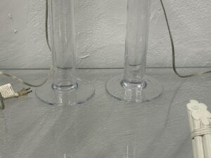 The Simon Pearce Glass Column Lamps, Pair, sit on a glass shelf with electrical cords and wall plug visible against a textured paint backdrop.