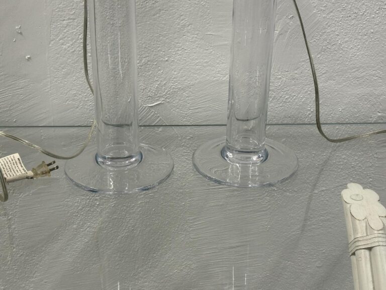 The Simon Pearce Glass Column Lamps, Pair, sit on a glass shelf with electrical cords and wall plug visible against a textured paint backdrop.