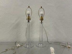 The Simon Pearce Glass Column Lamps, a pair, feature clear glass bodies with brass fittings and visible cords and plugs on a reflective surface, complete with bulbs.