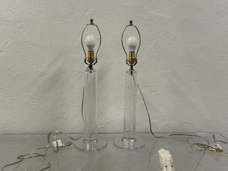 The Simon Pearce Glass Column Lamps, a pair, feature clear glass bodies with brass fittings and visible cords and plugs on a reflective surface, complete with bulbs.