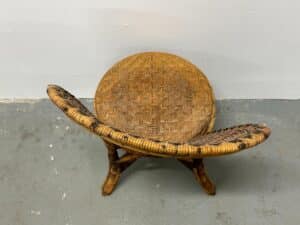 A brown, woven rattan stool with a curved backrest and crossed legs on a gray floor.