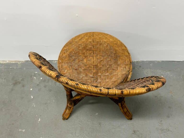 A brown, woven rattan stool with a curved backrest and crossed legs on a gray floor.