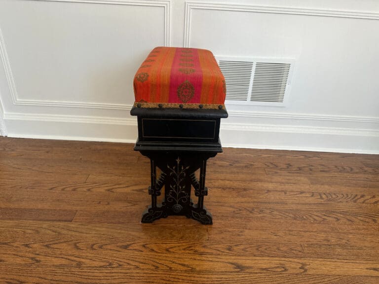 A Wood Carved Storage Bench featuring a red patterned cushion is positioned on a wooden floor next to a white wall with a vent.