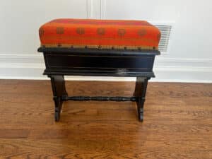 The Wood Carved Storage Bench features a vintage design with a black wooden body, an orange-red upholstered cushion, and rests on a wooden floor.