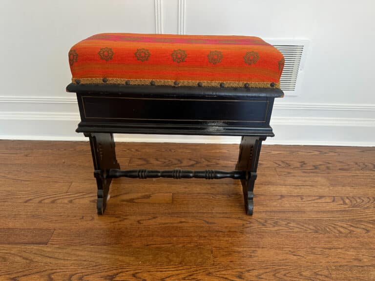 The Wood Carved Storage Bench features a vintage design with a black wooden body, an orange-red upholstered cushion, and rests on a wooden floor.