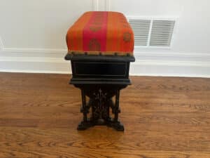 A Wood Carved Storage Bench with a red patterned fabric top sits on a wooden floor near a white wall with a vent.