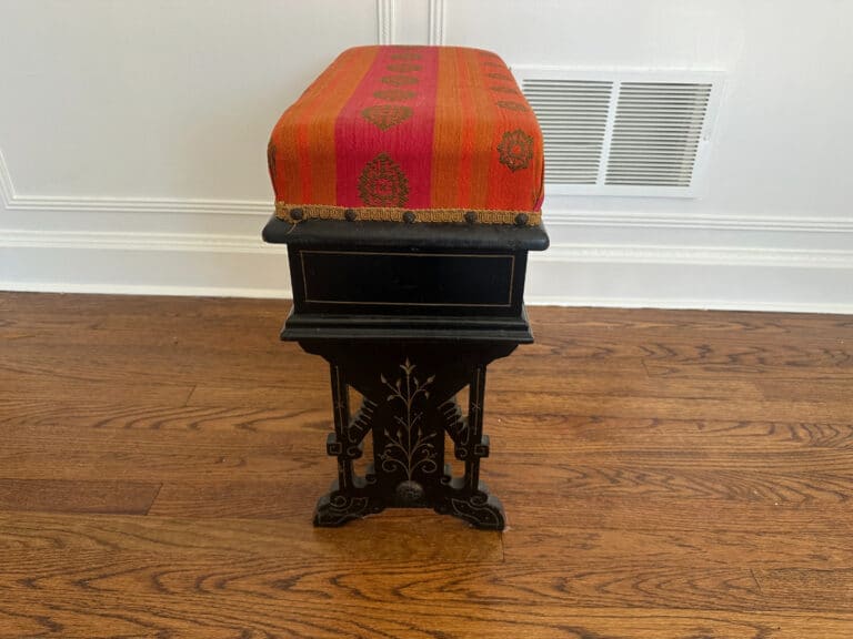 A Wood Carved Storage Bench with a red patterned fabric top sits on a wooden floor near a white wall with a vent.