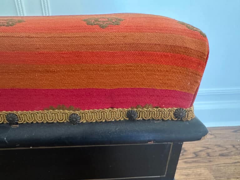 Close-up of a Wood Carved Storage Bench in red and orange stripes with decorative trim, positioned on a wooden floor beside a blue wall.