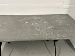 A weathered, rectangular metal table with a textured surface stands against a pale wall.