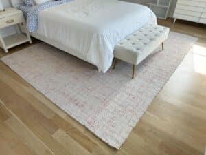 A white bed with a bench sits on a large patterned rug over light wood floors in a bright bedroom.