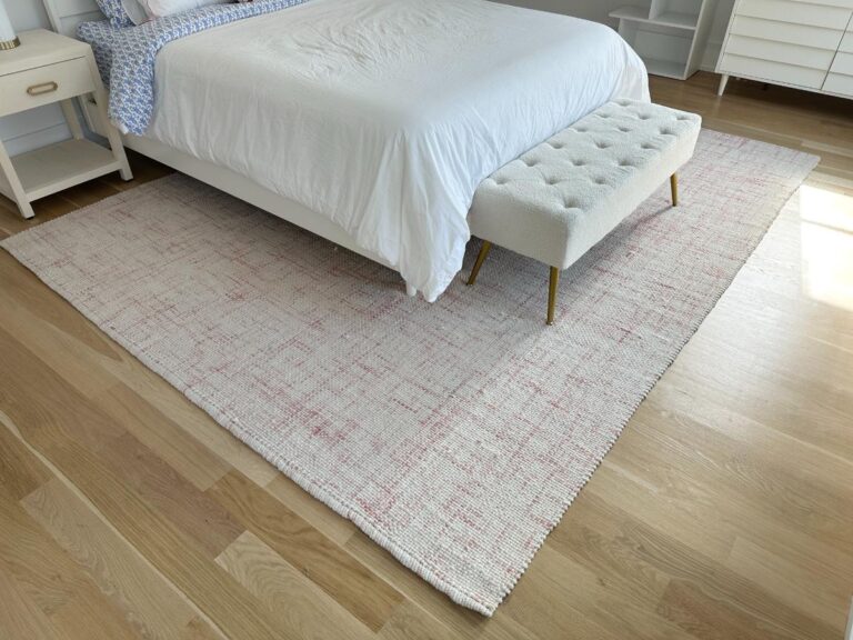 A white bed with a bench sits on a large patterned rug over light wood floors in a bright bedroom.