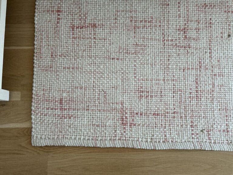 A close-up of a woven rug with a pink and cream pattern on a wooden floor.